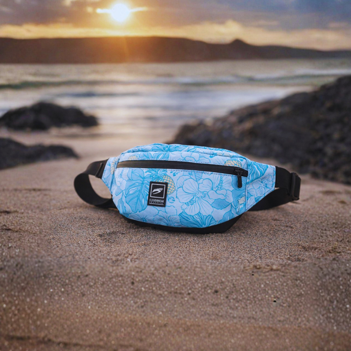 Blue floral-patterned waist bag on a sandy beach with a sunset in the background
