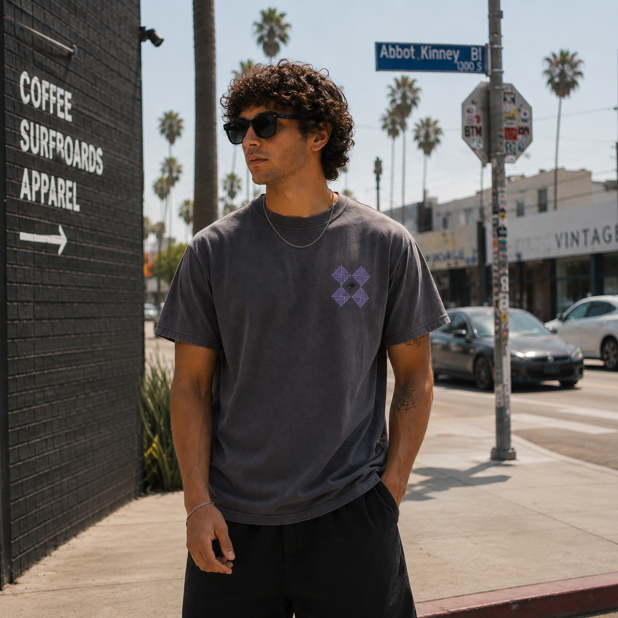 Man wearing sunglasses and a dark t-shirt with a logo, standing on a sidewalk with palm trees and a street sign in the background.