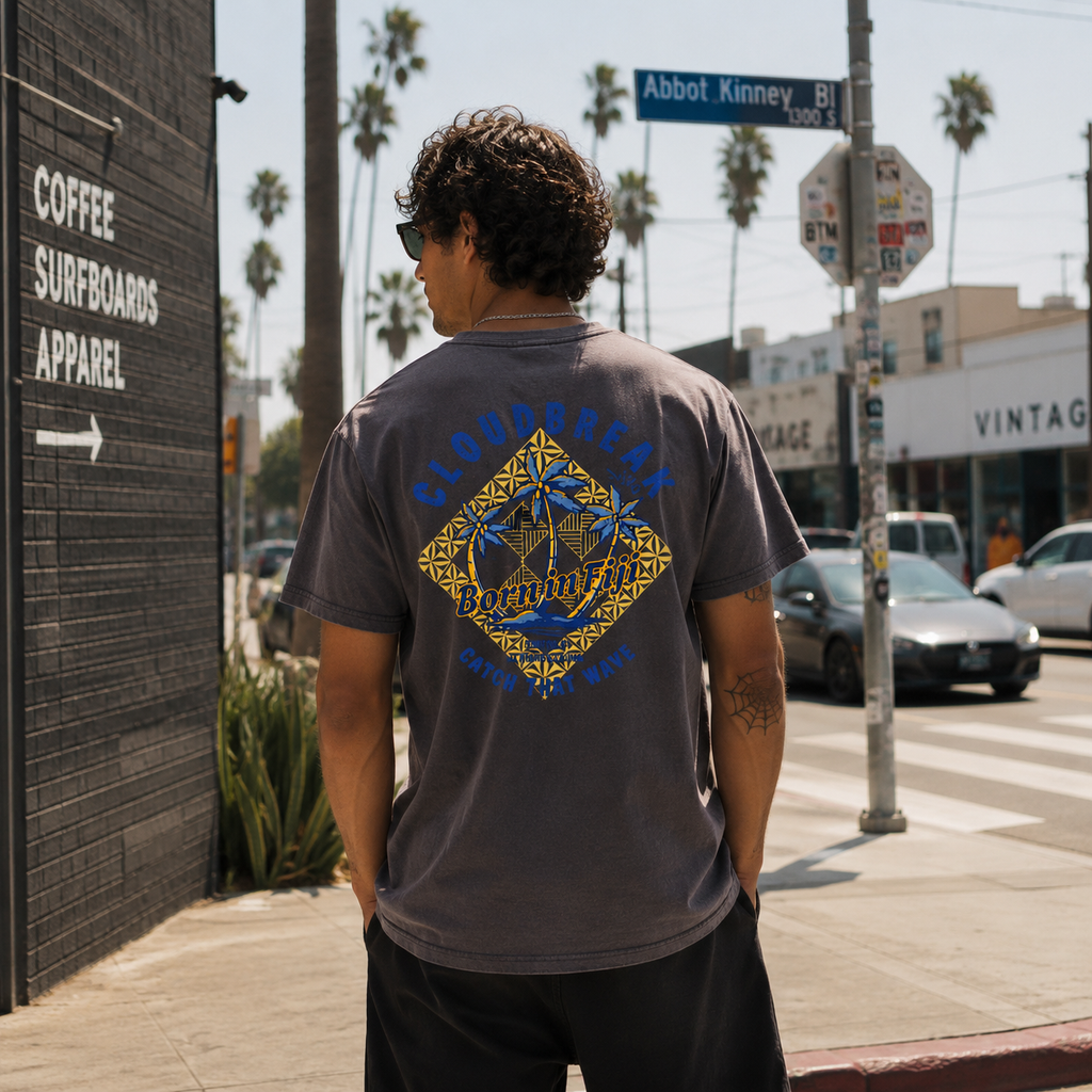 Man wearing a gray t-shirt with a colorful design on the back, standing on a sidewalk with palm trees and buildings in the background.